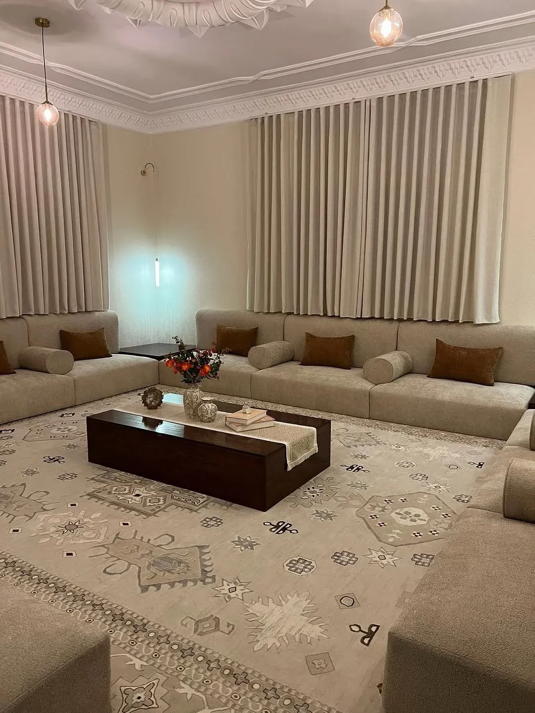 مجلس عربي مودرن بيج منخفض على ثلاث جهات بقطع منفصلة ومساند أسطوانية وطاولات خشب في الزوايا